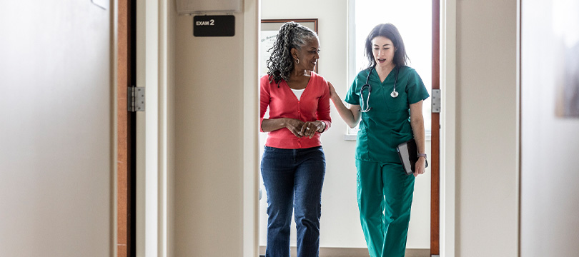 An image of a woman talking with her doctor while the walk in a doctor's office. The doctor's hand is on the patient's shoulder.