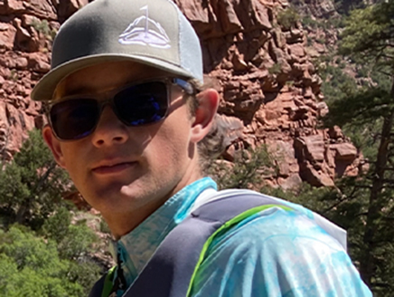 An image of Talmage wearing a hat and sunglasses while standing in front of cliffs on a hike.