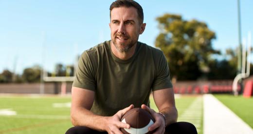 An image of Alex Smith wearing a green shirt sitting on a field holding a football in his hands