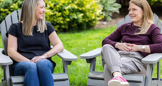 An image of two women sitting on Adirondack chairs outside with plants in the background.