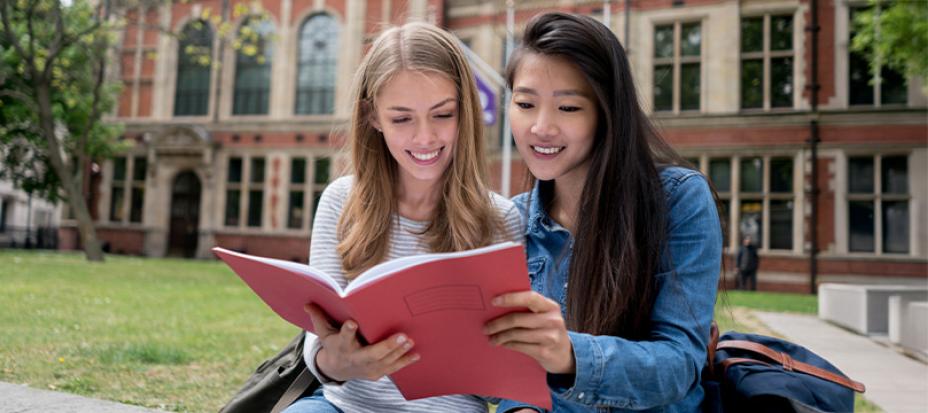 An image of two women sitting on the ground while reading a red book together. There is a red brick building with many windows behind them.