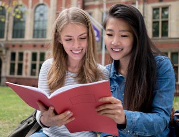 An image of two women sitting on the ground while reading a red book together. There is a red brick building with many windows behind them.