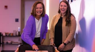An image of two women standing next to each other and laughing while playing a game of shuffleboard
