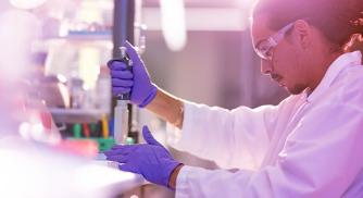 An image of a Vertex scientist in a lab wearing a white coat and purple gloves while using lab equipment during an experiment