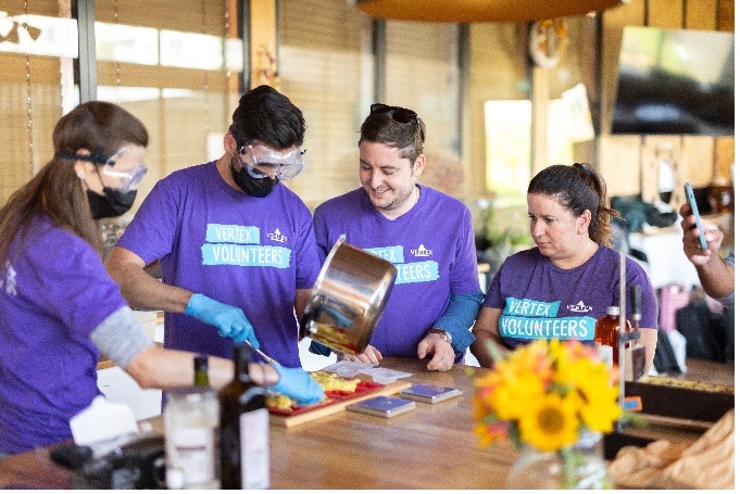 An image of four Vertexians wearing purple Vertex Volunteers shirts while combining ingredients from a pot onto a cutting board.