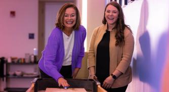 Two women standing next to each other and laughing while playing a game of shuffleboard