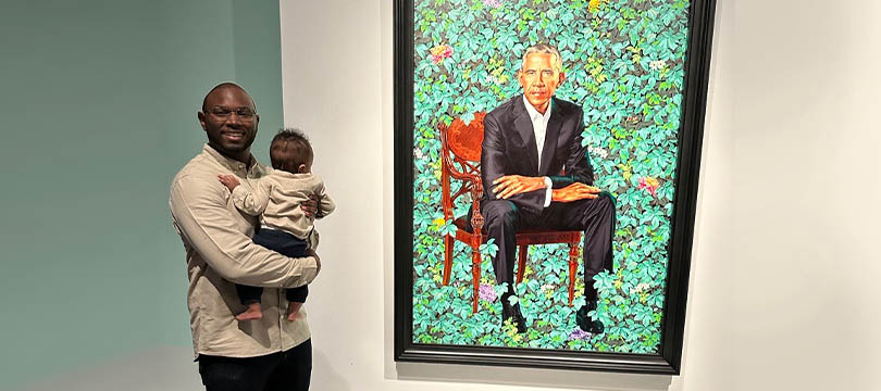 An image of Marcus Kernizan holding his young child while standing next to a painting of former U.S. President Barack Obama