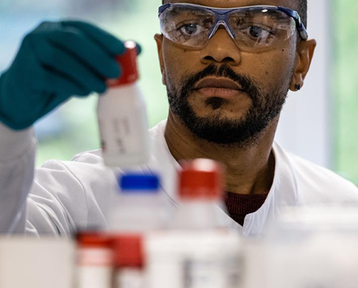 An image of a scientist in the Vertex Pharmaceuticals lab grabbing a bottle. He is wearing goggles, a lab coat and gloves