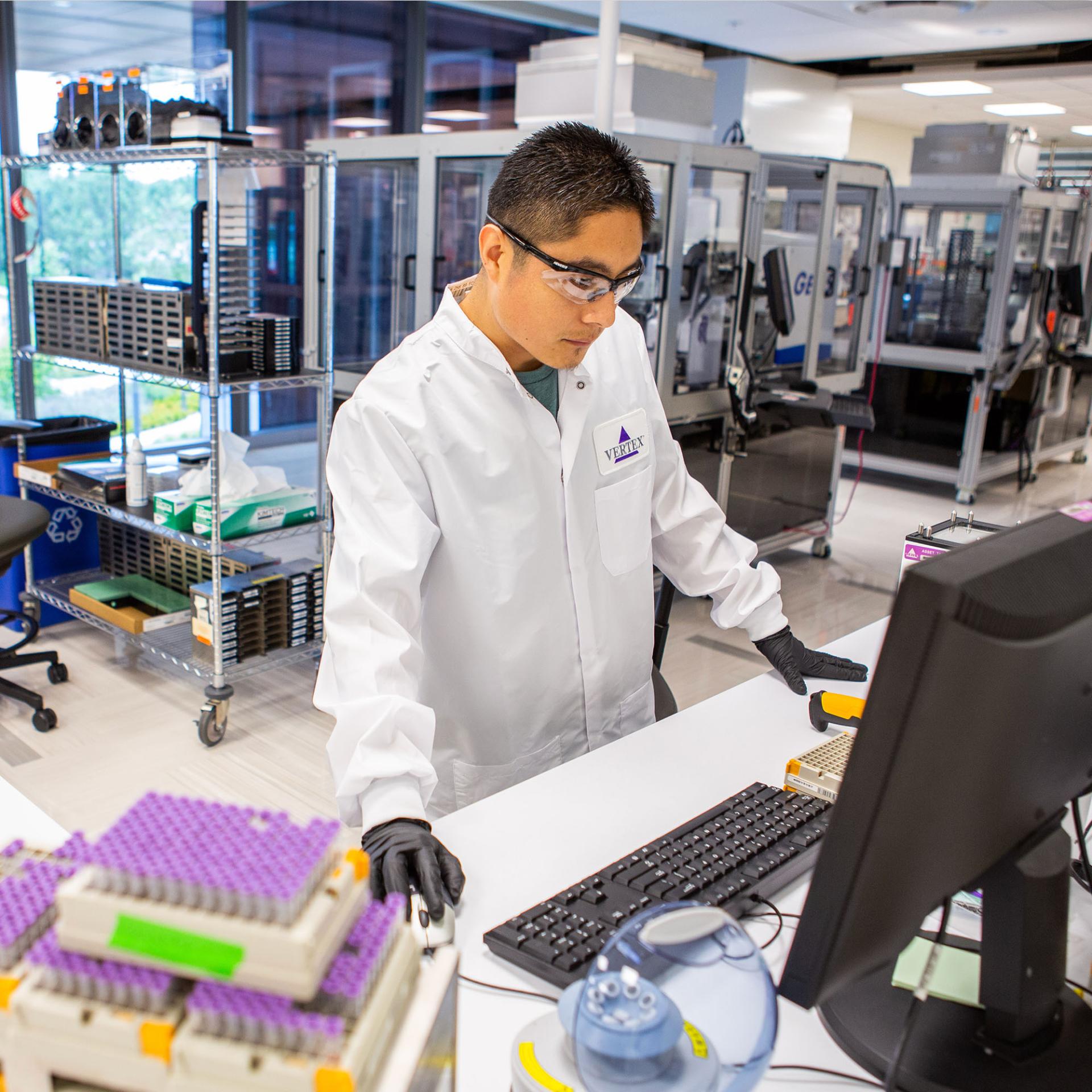 A Vertex scientist in a white lab coat standing at a computer using a mouse