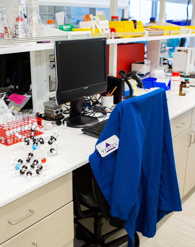 An image of a Vertex lab with a computer screen and a chair with a blue lab coat resting on the back. There are vials, beakers, and molecule models on the table next to the computer screen.