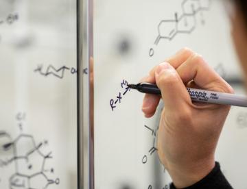 A closeup of a hand writing a formula on glass