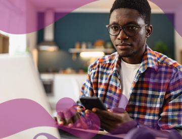 An image of a man looking at a computer screen with a phone in his hand with a purple watercolor effect on the outside of the image