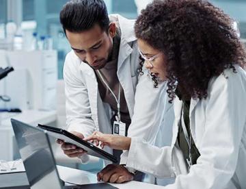 An image of two scientists in lab coats who are standing in a lab looking at a tablet.
