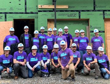 An image of a group of Vertexians participating in Day of Service, wearing purple Day of Service shirts and hard hats, with a house in the midst of construction behind them