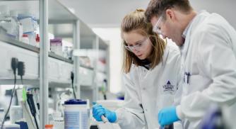 An image of two Vertex scientists wearing white lab coats and blue gloves working together in the lab on an experiment