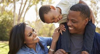 An image of a child on top of his father's shoulders while his mother stands next to them both while smiling