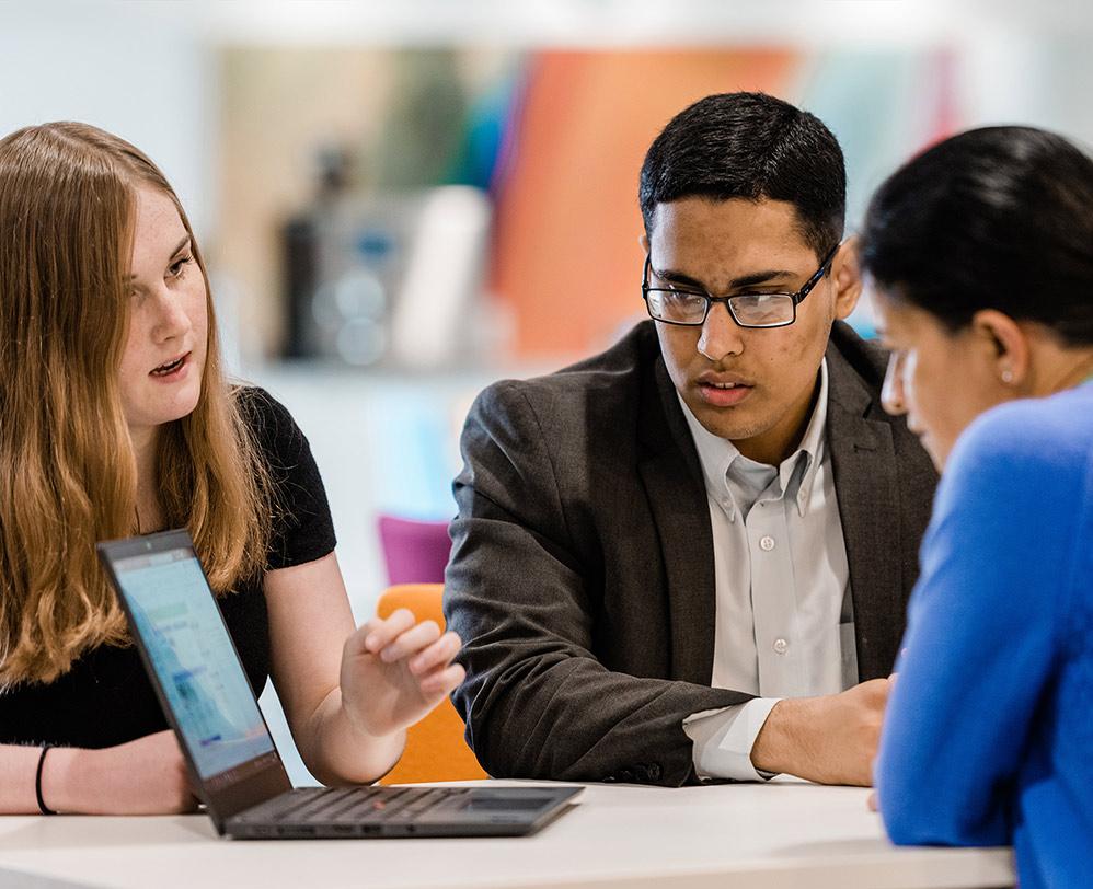 An image of two Year 12 students in Vertex Pharmaceuticals' Oxford, U.K., Research Site sitting at a table, looking at a laptop, and having a conversation with a female research scientist.
