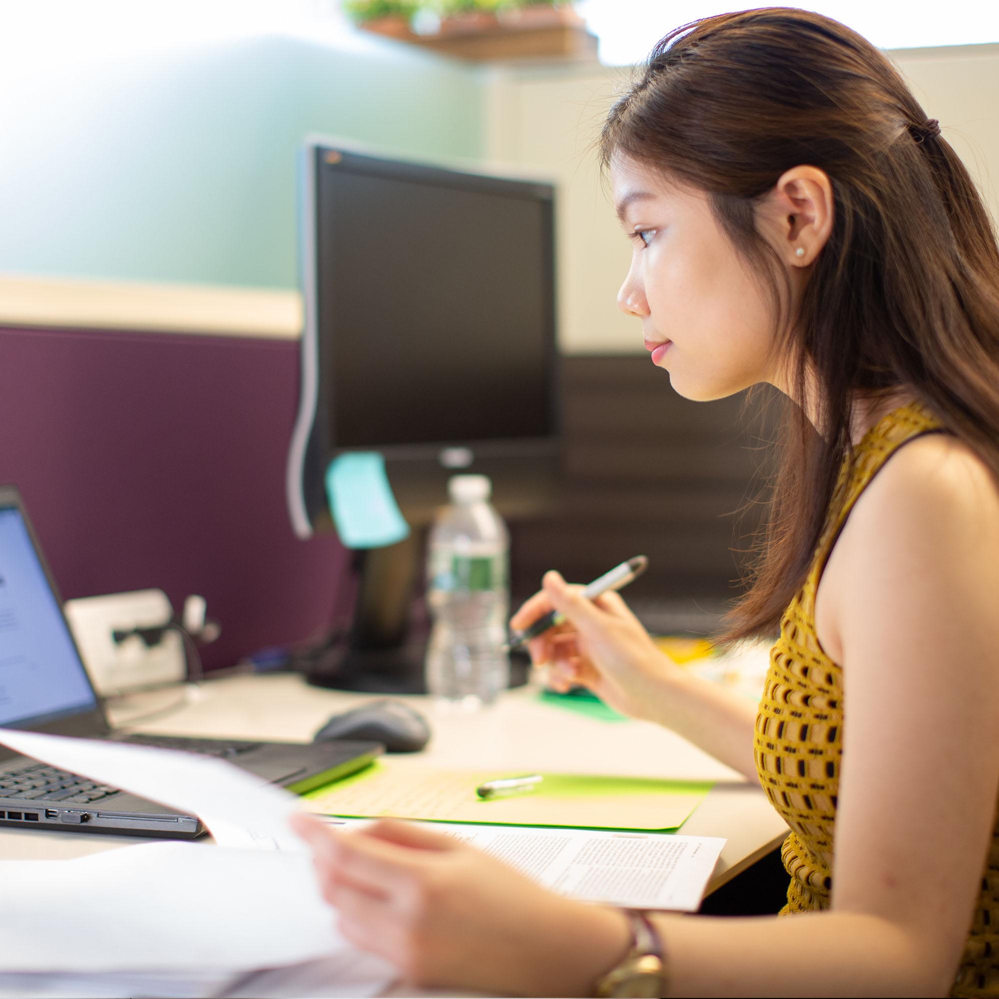 An intern uses a laptop at her desk while taking notes