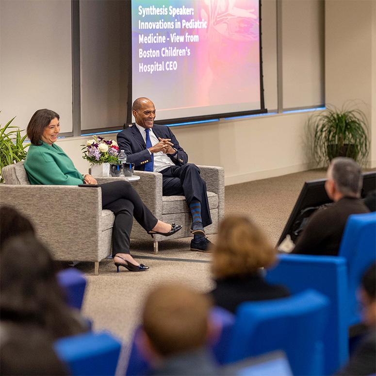 An image of Vertex Pharmaceuticals CEO Reshma Kewalramani and a guest speaker laughing while interacting with the crowd during a Vertex speaking event