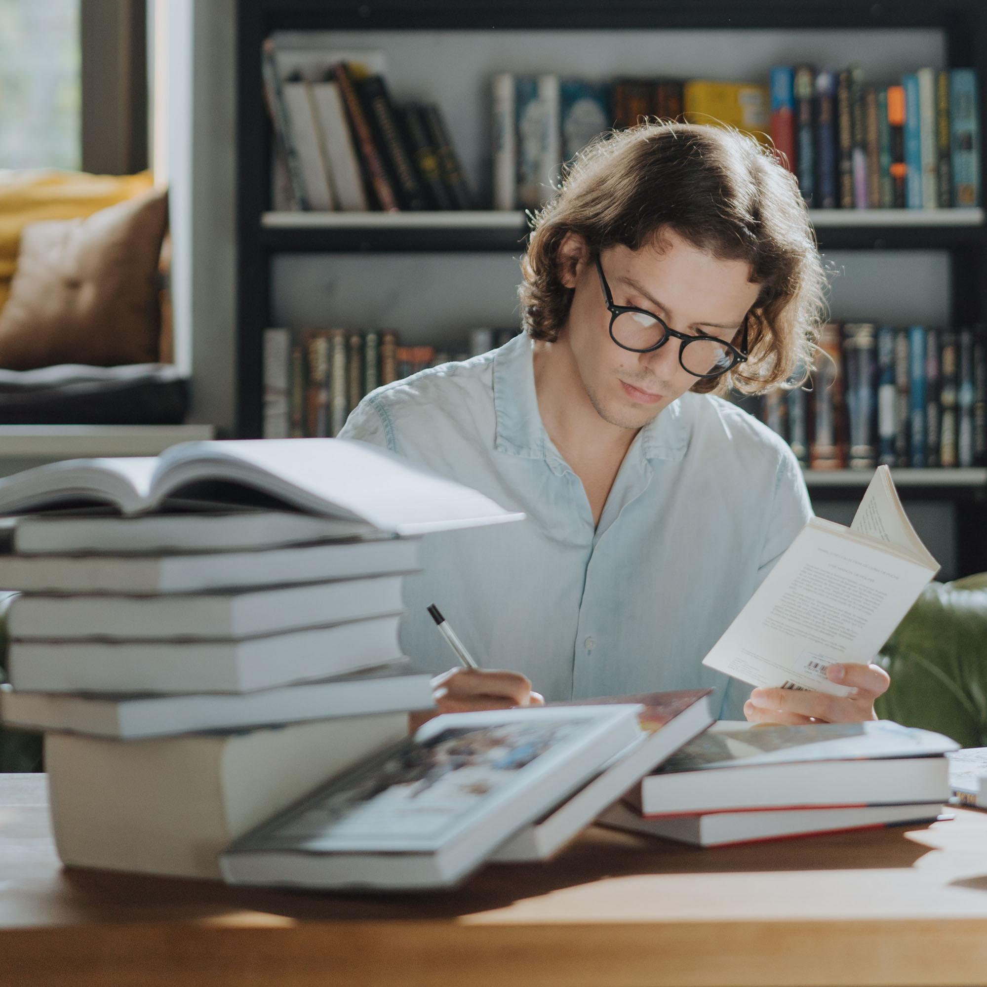 A man wearing glasses takes notes while reading a book