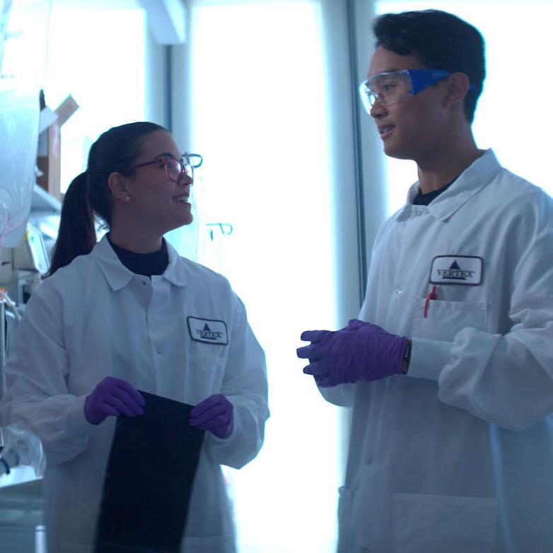 An image of two Vertex scientists wearing white lab coats conversing while surrounded by lab technology