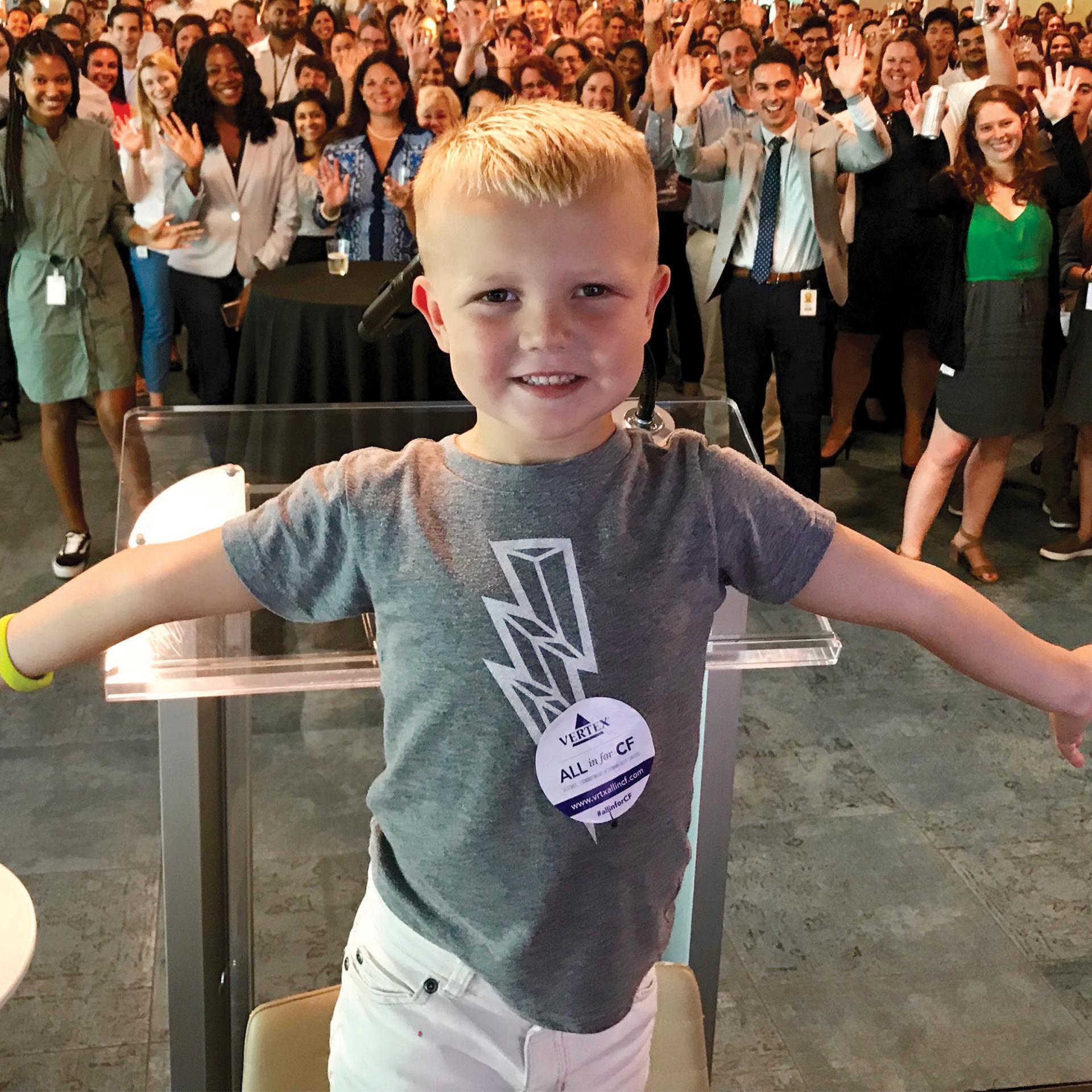 An image of a boy standing in front of a crowd of Vertex employees