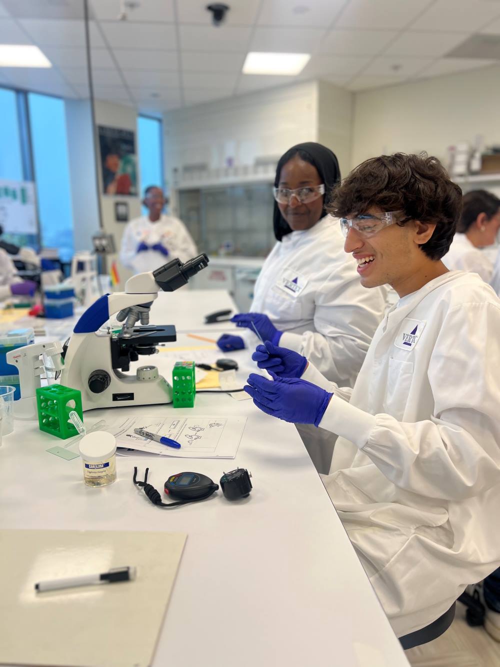 An image of two students in a Vertex Learning Lab wearing white Vertex lab coats while working on an experiment.