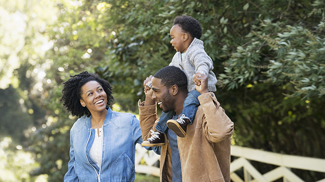 An image of a family with the mother smiling at the child who is sitting on his father's shoulders