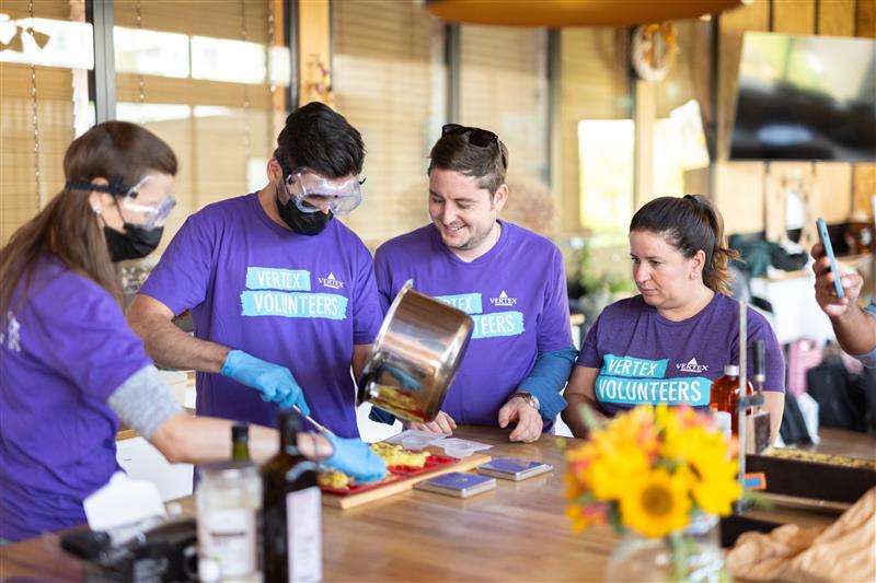 An image of four Vertexians wearing purple Vertex Volunteers shirts while combining ingredients from a pot onto a cutting board.