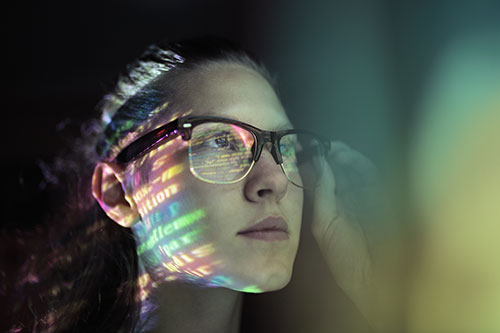 An image of a woman with glasses with a rainbow colored reflection shining on her face
