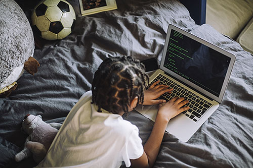 An image of a student laying in bed with a soccer ball on it while working on a laptop