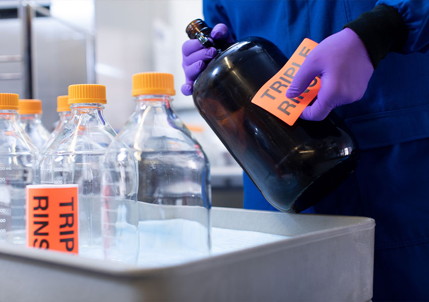 An image of a scientist in blue scrubs and wearing purple gloves applying a label that reads “TRIPLE RINSE” to a bottle in a lab.