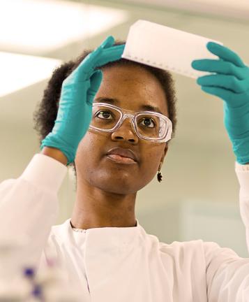 A scientist wearing goggles, a lab coat and gloves holds up a tray to examine
