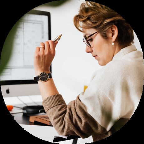 A woman sitting at a computer holding a pen and looking down at papers