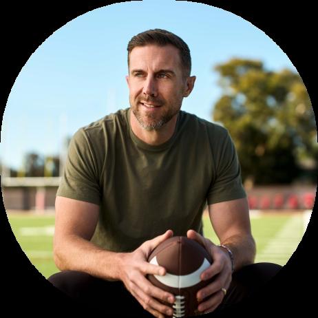 An image of Alex Smith sitting on a football field holding a football in his hands while wearing a green short sleeve shirt