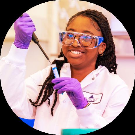 A Black female high school student conducting an experiment in Vertex Pharmaceuticals' Boston Learning Lab. She is wearing a lab coat, safety glasses and gloves, and holding a pipette.