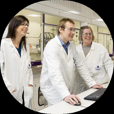 3 Vertex employees in lab coats standing at a computer in the lab