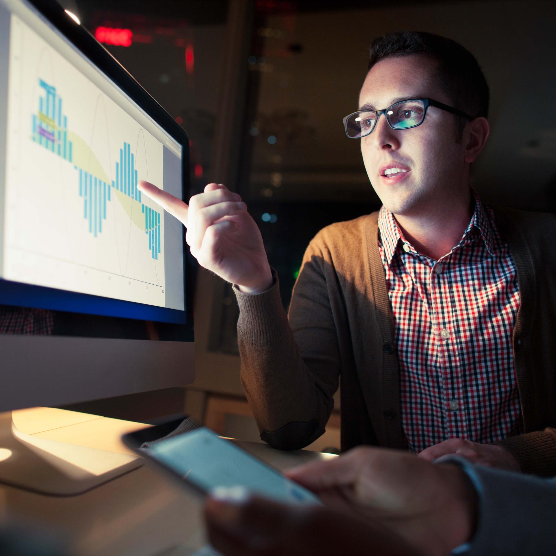 An image of a man wearing glasses pointing at a bar graph on a monitor