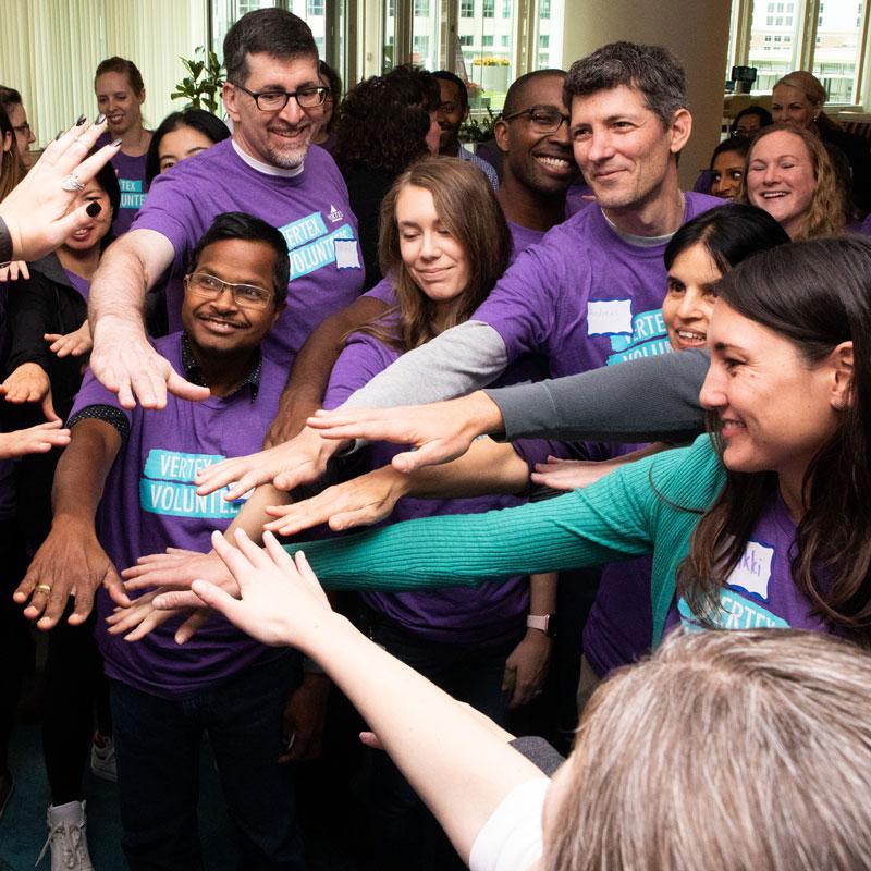 An image of a large group of Vertexians wearing purple Vertex Volunteers shirts all putting their hands together