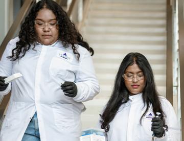 An image of two female scientists wearing white Vertex lab coats and walking down a set of stairs