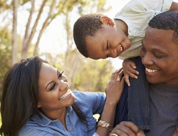 An image of a child on top of his father's shoulders while his mother stands next to them both while smiling