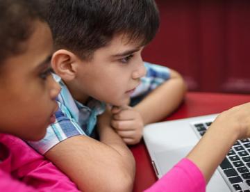 An image of a girl and boy pointing to the screen of an open laptop