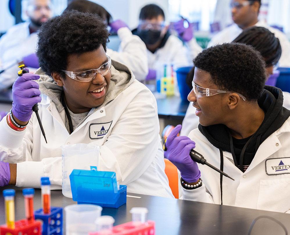An image of two Black male high school students conducting an experiment in Vertex Pharmaceuticals' Boston Learning Lab. They are both wearing a lab coat, safety glasses and gloves, and they are each holding a pipette.
