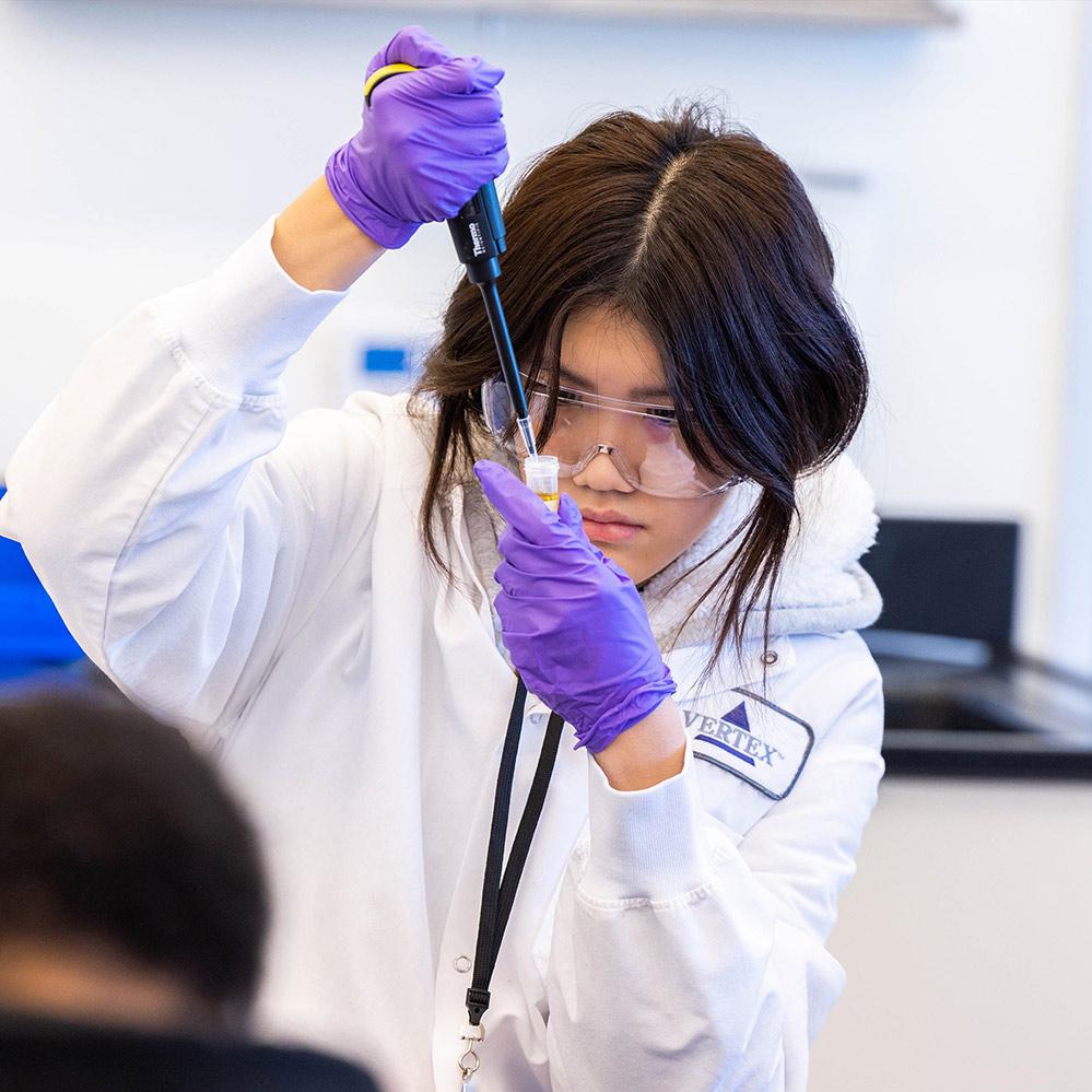 An image of a female high school student conducting an experiment in Vertex Pharmaceuticals' Boston Learning Lab. She is wearing a lab coat, safety glasses and gloves, and practicing pipetting technique.