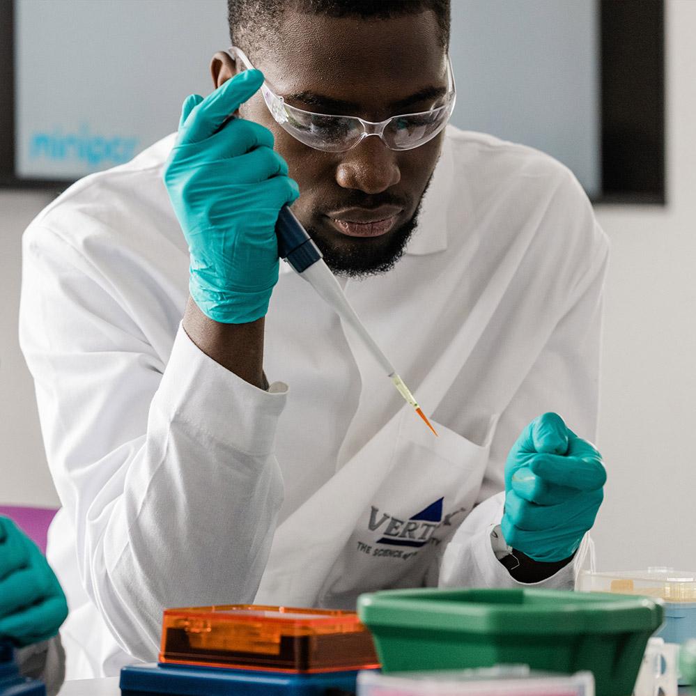 An image of a Black, male student conducting an experiment in Vertex Pharmaceuticals' Oxford, U.K., Learning Lab. He is wearing a lab coat, safety classes and gloves, and practicing pipetting technique.