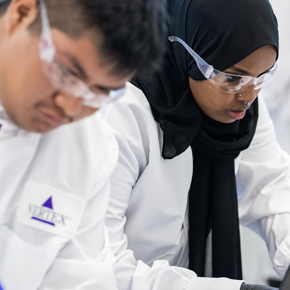 An image of two high school students (one female, one male) conducting an experiment in Vertex Pharmaceuticals' San Diego Learning Lab. They are both wearing lab coats and safety glasses.