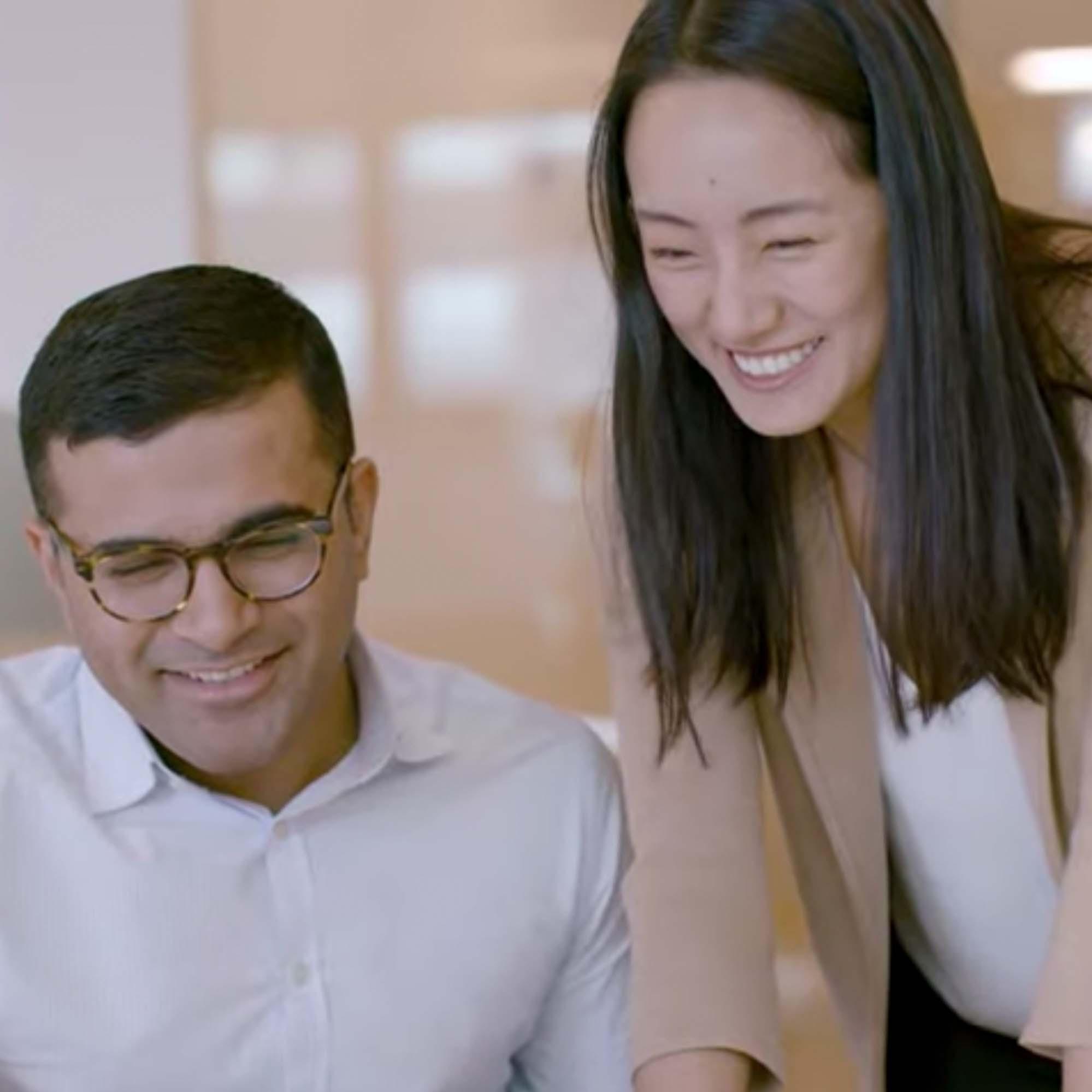 A man sitting down smiling with a smiling female colleague standing next to him