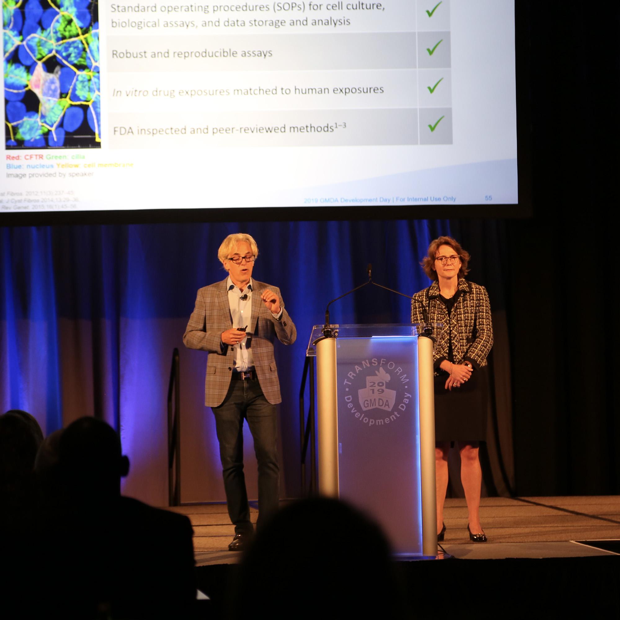 2 presenters on stage reviewing a scientific slide