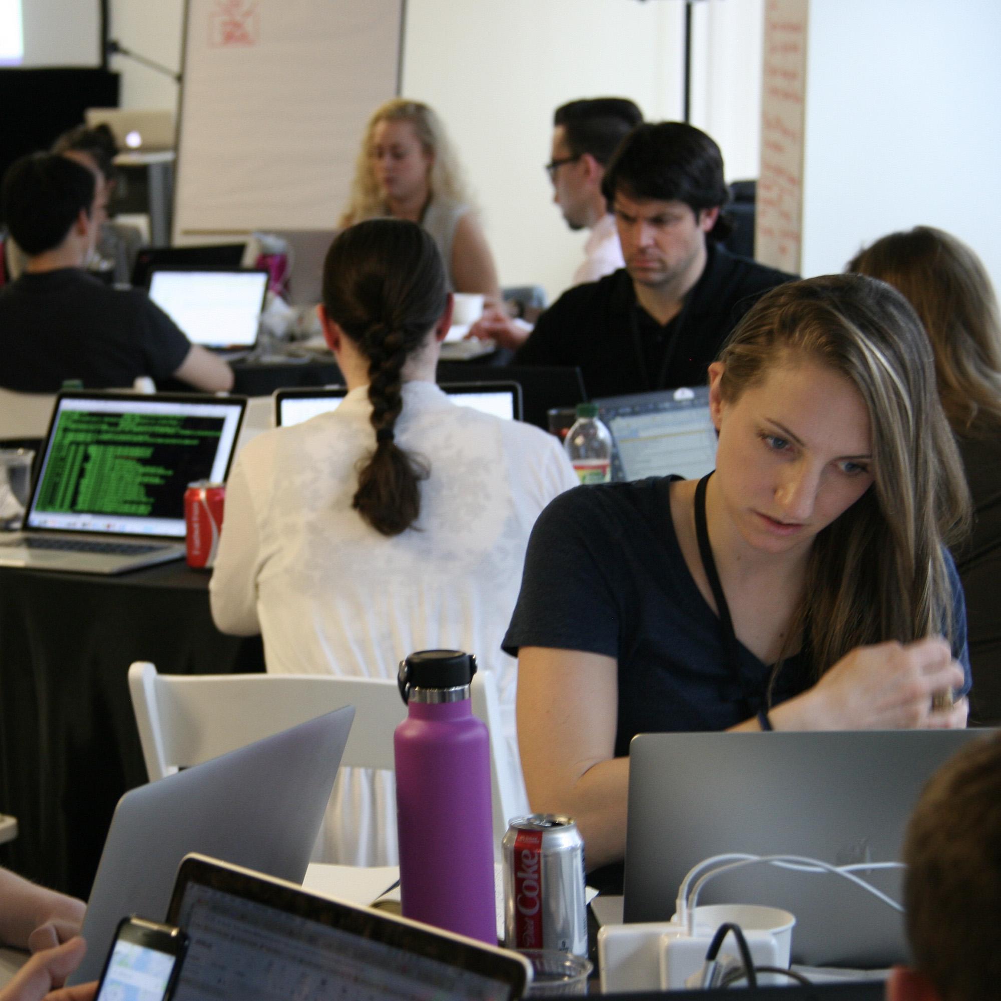 Participants in a hackathon sit at tables working on their laptops