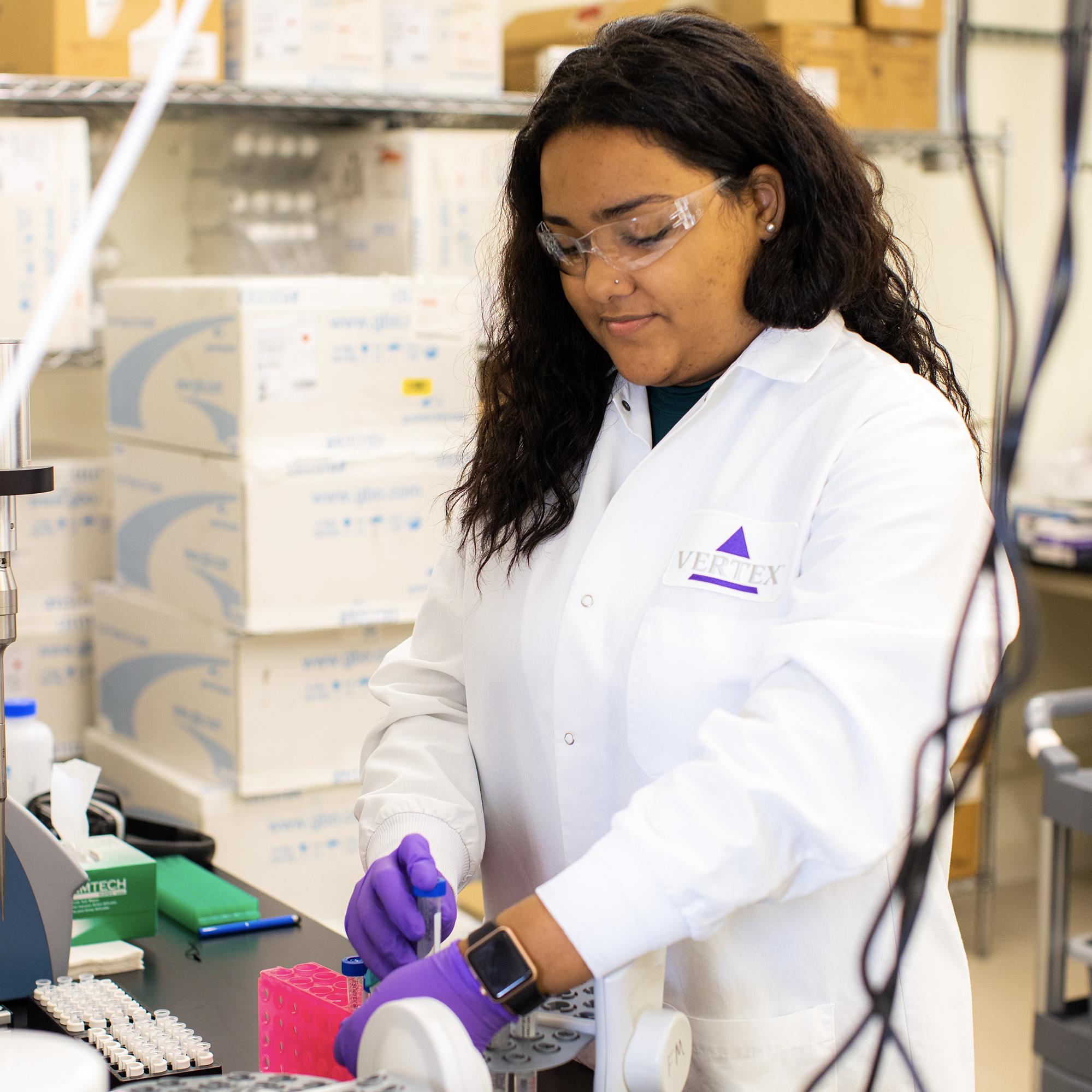 An image of a female high school student conducting an experiment in Vertex Pharmaceuticals' Boston Learning Lab. She is wearing a lab coat, safety glasses and gloves, and standing at a lab bench.